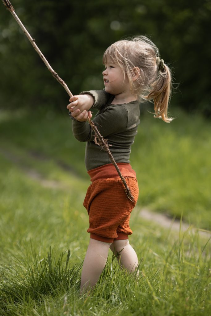 Ein etwa dreijähriges Mädchen mit einer rostroten, kurzen Musselinhose spielt mit einem Stock